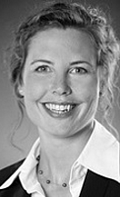 Porträt einer lächelnden Frau mit lockigem Haar, die eine Bluse und einen Blazer trägt. Sie schaut direkt in die Kamera und strahlt Freundlichkeit und Professionalität aus. Der Hintergrund ist unscharf und in Grau gehalten.